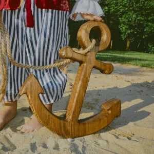Drewniana kotwica dekoracyjna na piasku, obok osoba w stroju marynarza.