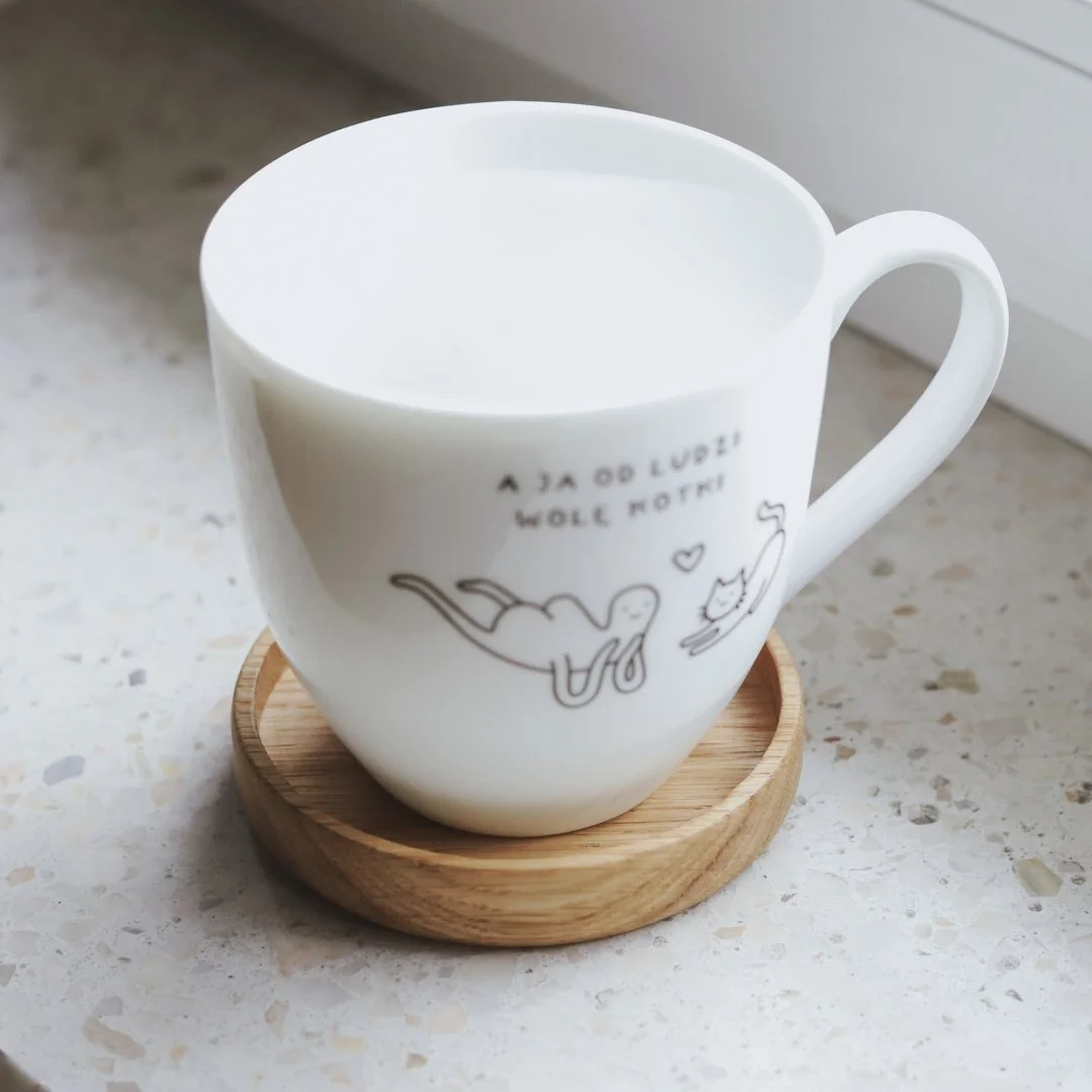 Biały kubek z grafiką na drewnianej podkładce.