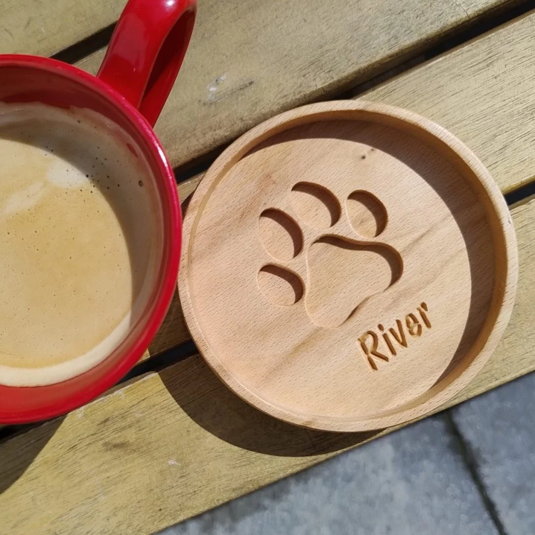 Drewniana podkładka z wyciętym śladem łapy i napisem River obok czerwonego kubka z kawą.